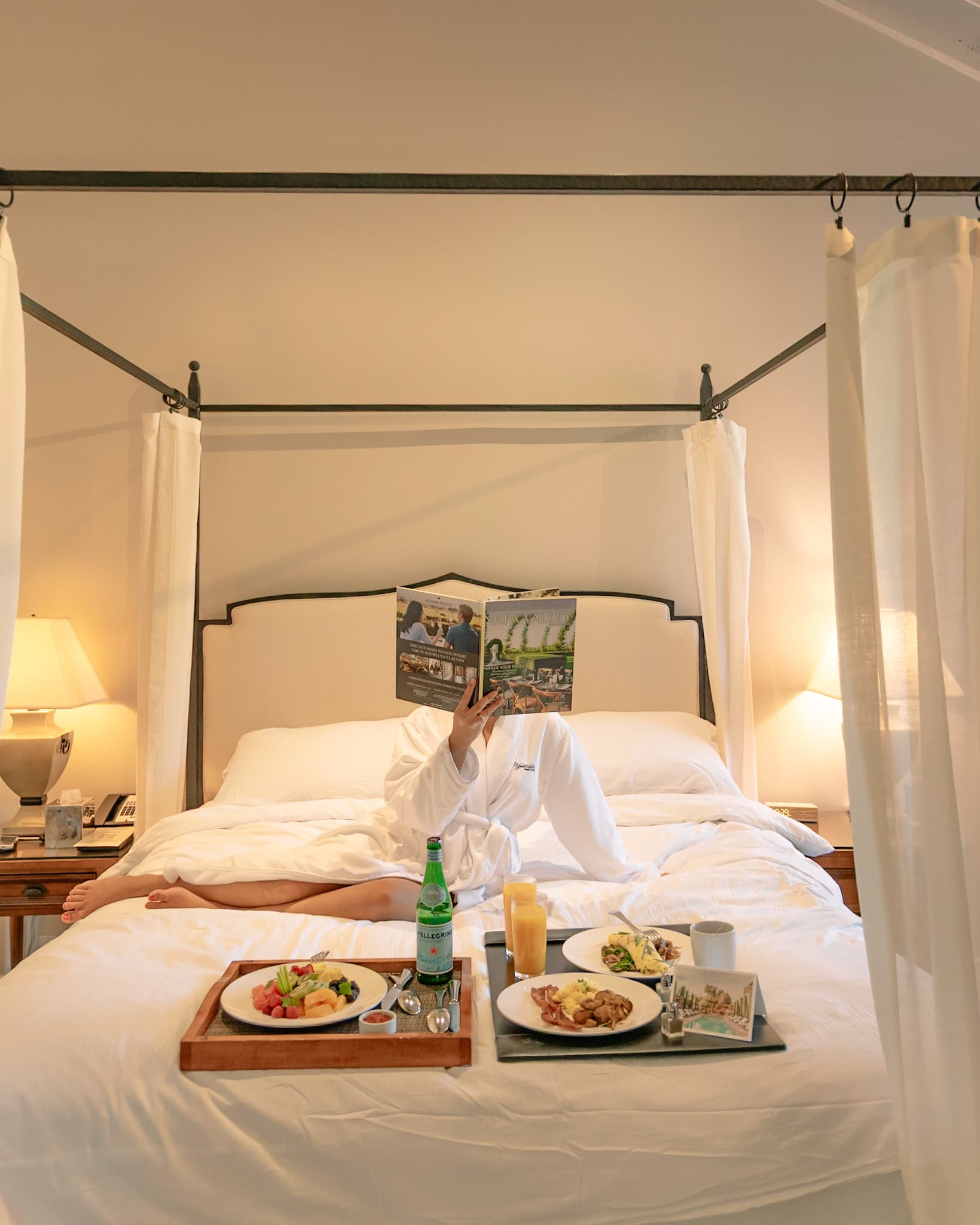 Tram Le in a white robe reading in bed with breakfast trays inside a canopy guest room at Hotel Yountville in Napa Valley.